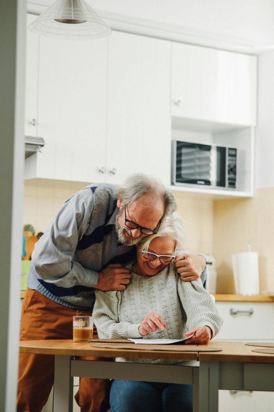 Two seniors at their kitchen looking at a paper, smiling.